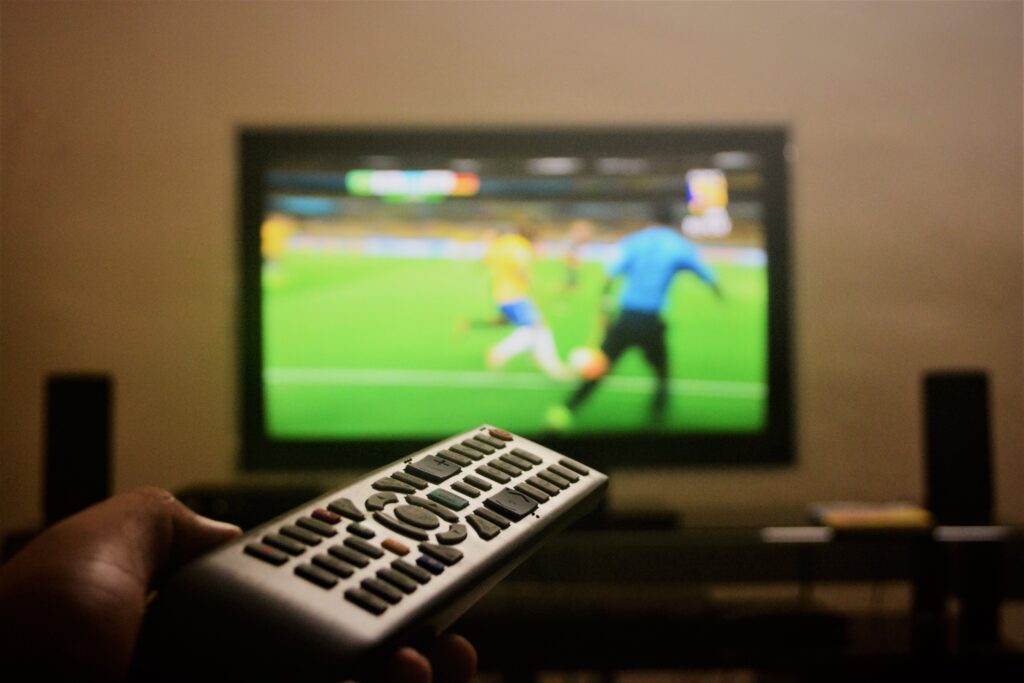 Viewer holding a remote while watching football on television, illustrating domestic broadcaster coverage on a large screen.